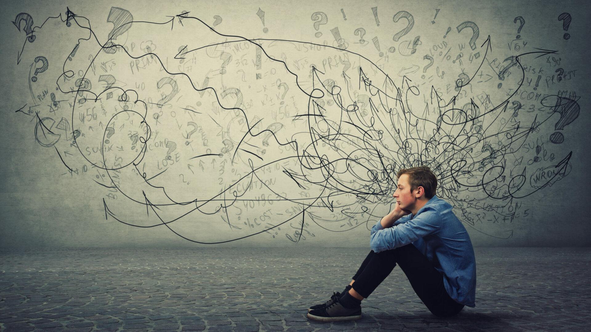 Picture of a man sitting sadly on the floor, seemingly overwhelmed by the questioning and depressive thoughts illustrated with scribbles