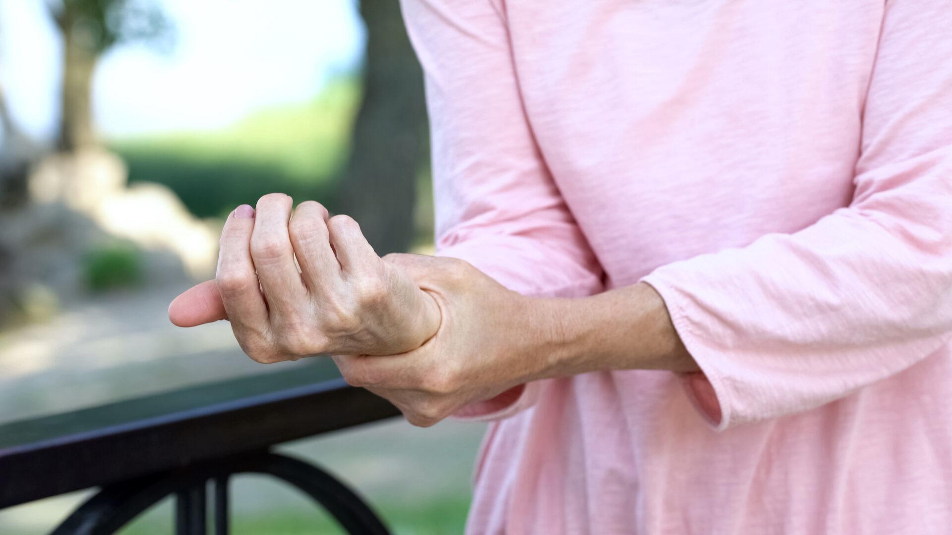 A close-up of a woman wearing a pink shirt, holding and massaging her wrist with a pained expression.