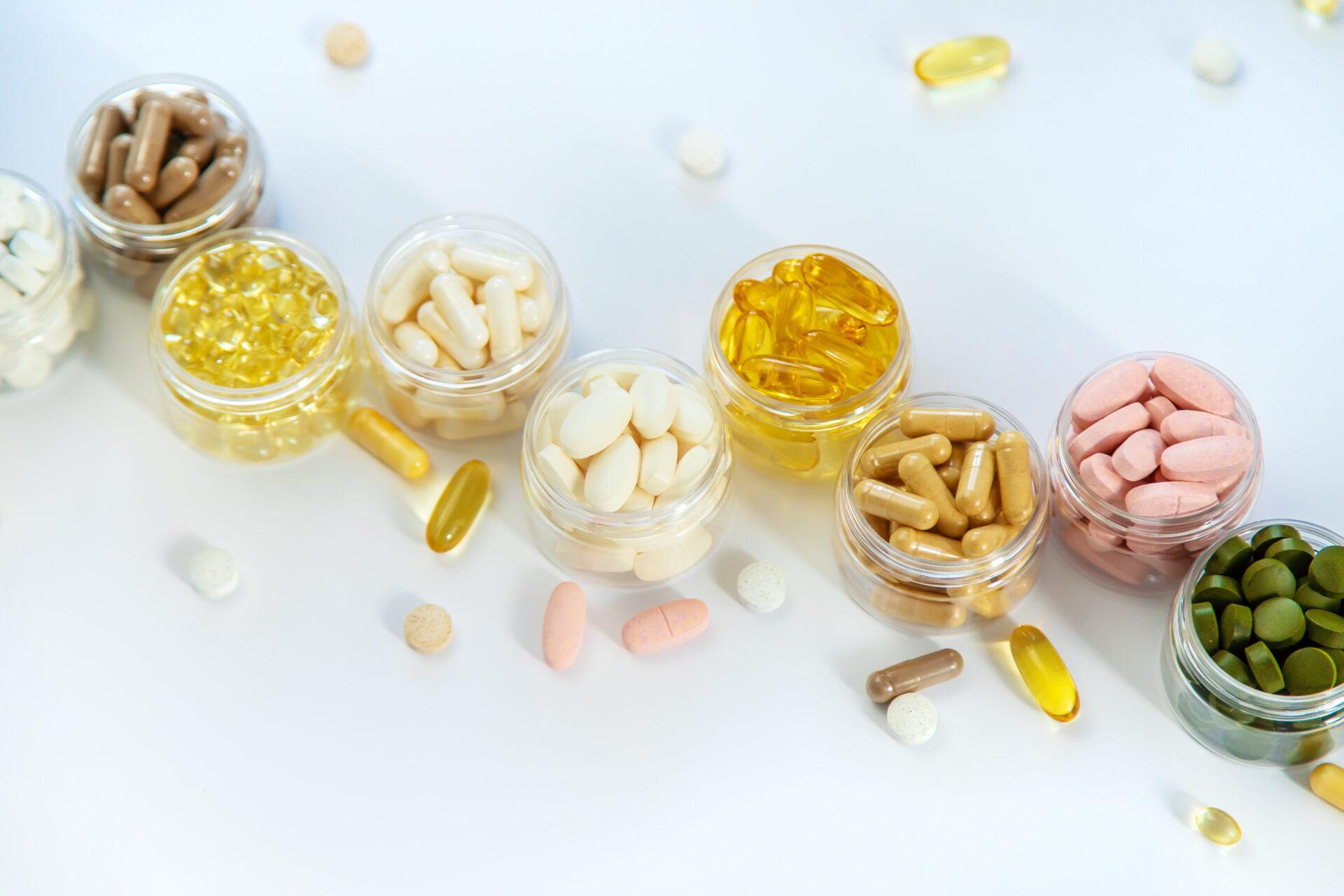 An assortment of various vitamins and supplements in small glass jars displaying different shapes, sizes, and colours of capsules and tablets.