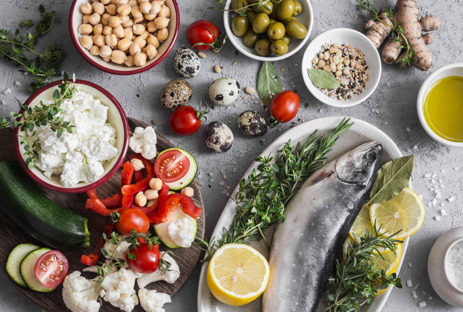 A variety of Mediterranean diet foods, including fresh fish, vegetables, chickpeas, quail eggs, olives, and herbs, arranged on a grey surface.