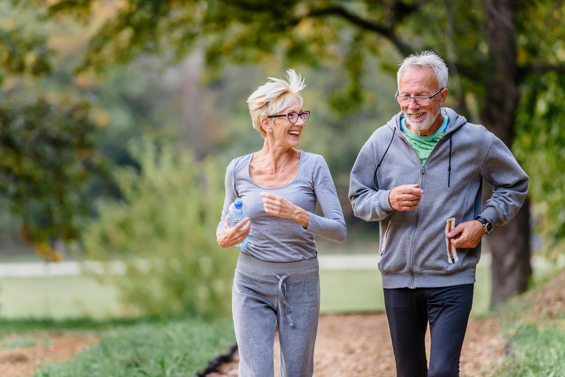 A senior couple dressed in athletic clothing, jogging and smiling together on a path in a park with trees in the background.