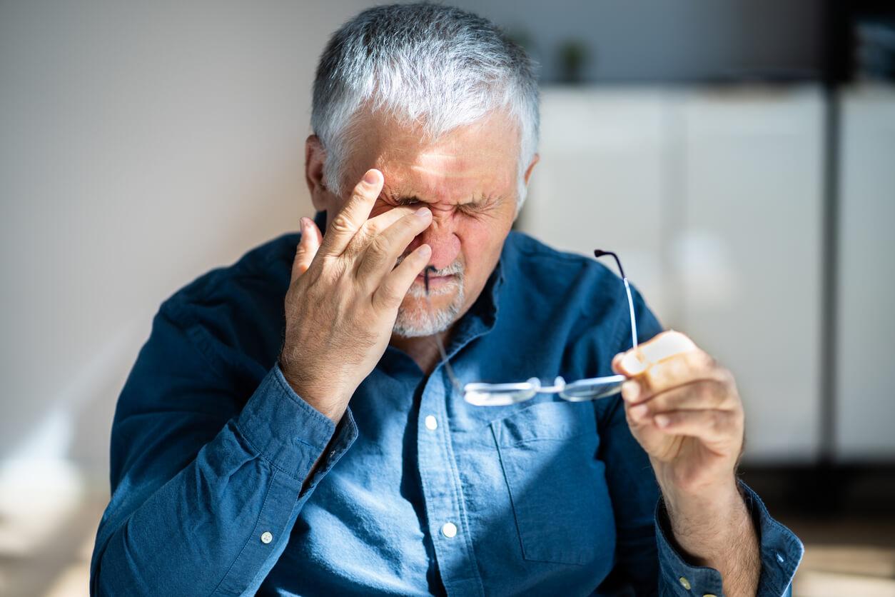 A senior man appears to be in discomfort as he rubs his eyes with one hand, holding a pair of glasses in the other.