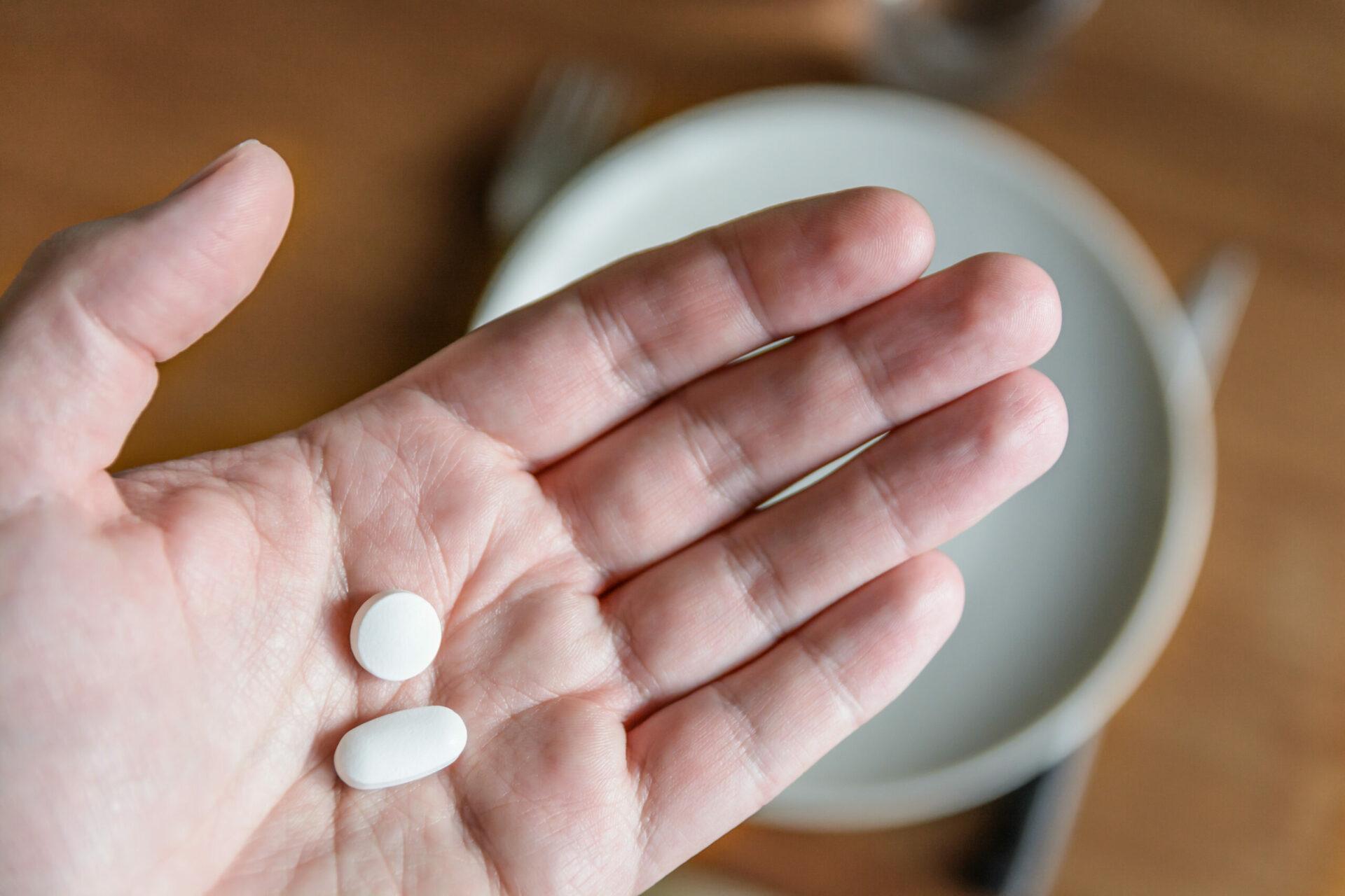 Close-up view of a male hand holding two white pills in the palm above a blurry background showing a dining table with plate