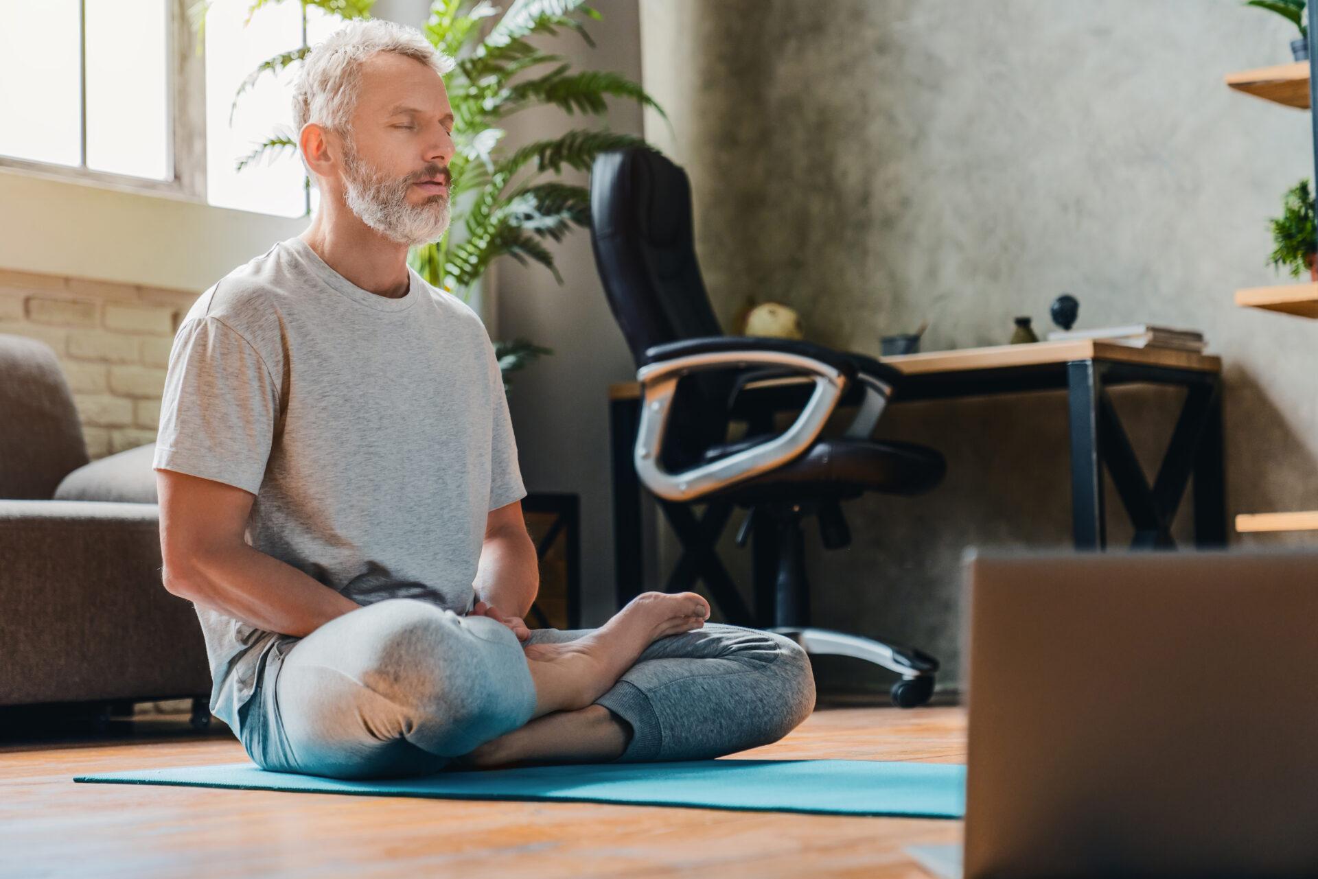 Middle-aged man meditating at home using laptop.