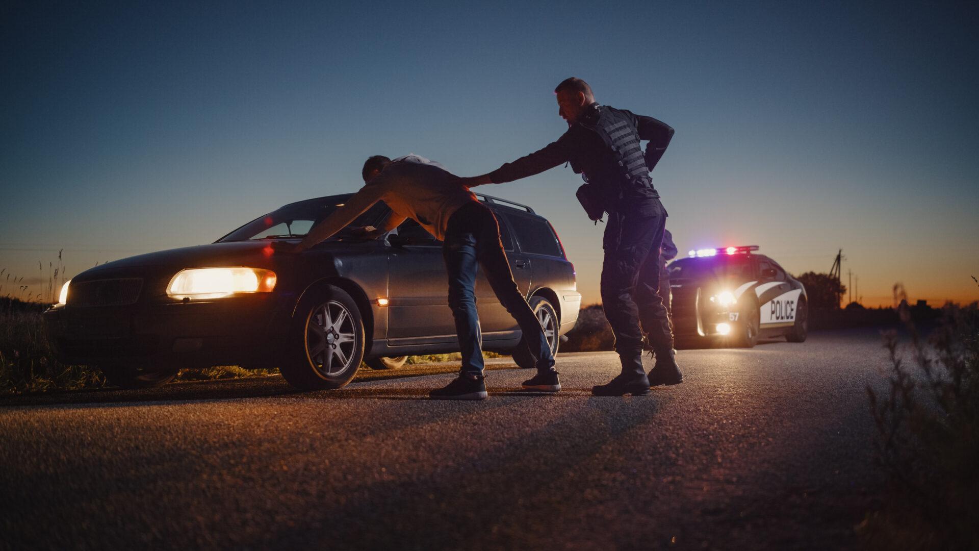 A law enforcement officer performing a pat-down on a driver who is standing with his hands on the car's hood.