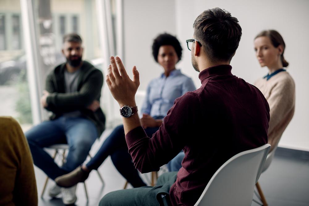  Rear view of man with raised hand on group therapy in a medical center