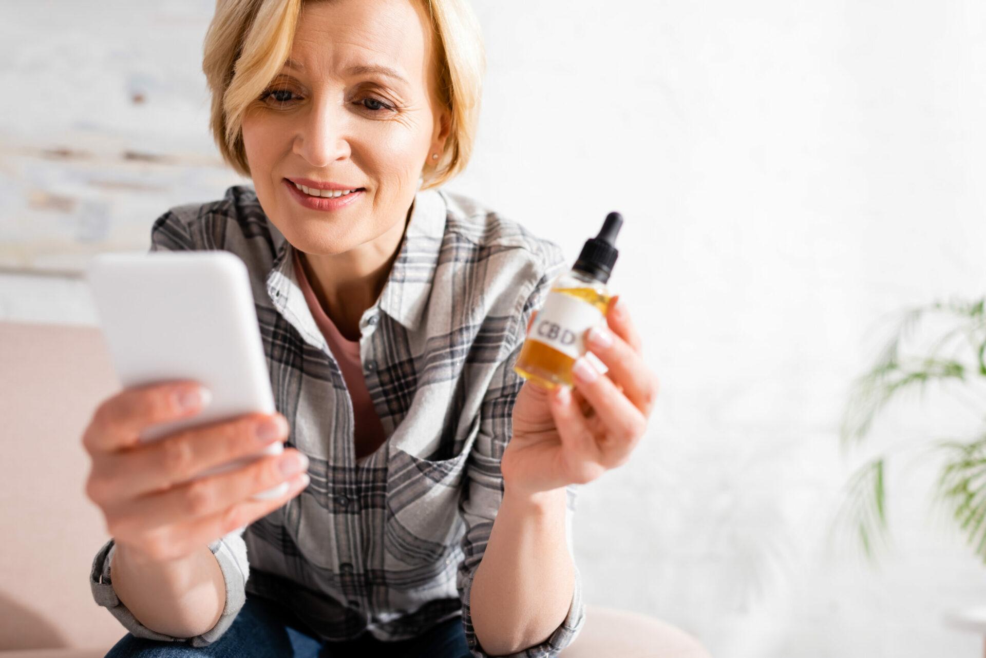 Image of a happy woman holding a bottle of CBD oil and pressing her phone. 