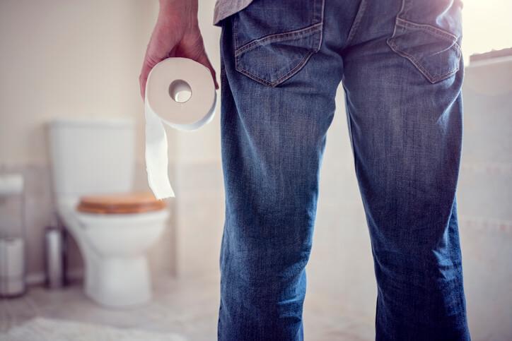 A man in a bathroom holds a roll of toilet paper, standing next to a toilet, emphasising a common household situation.