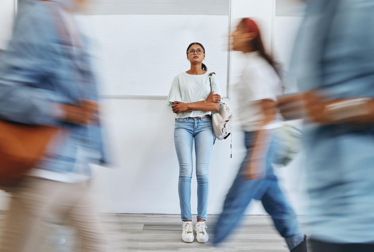A young woman stands alone in a bustling corridor, surrounded by blurred figures in motion, symbolising isolation amidst a crowd.