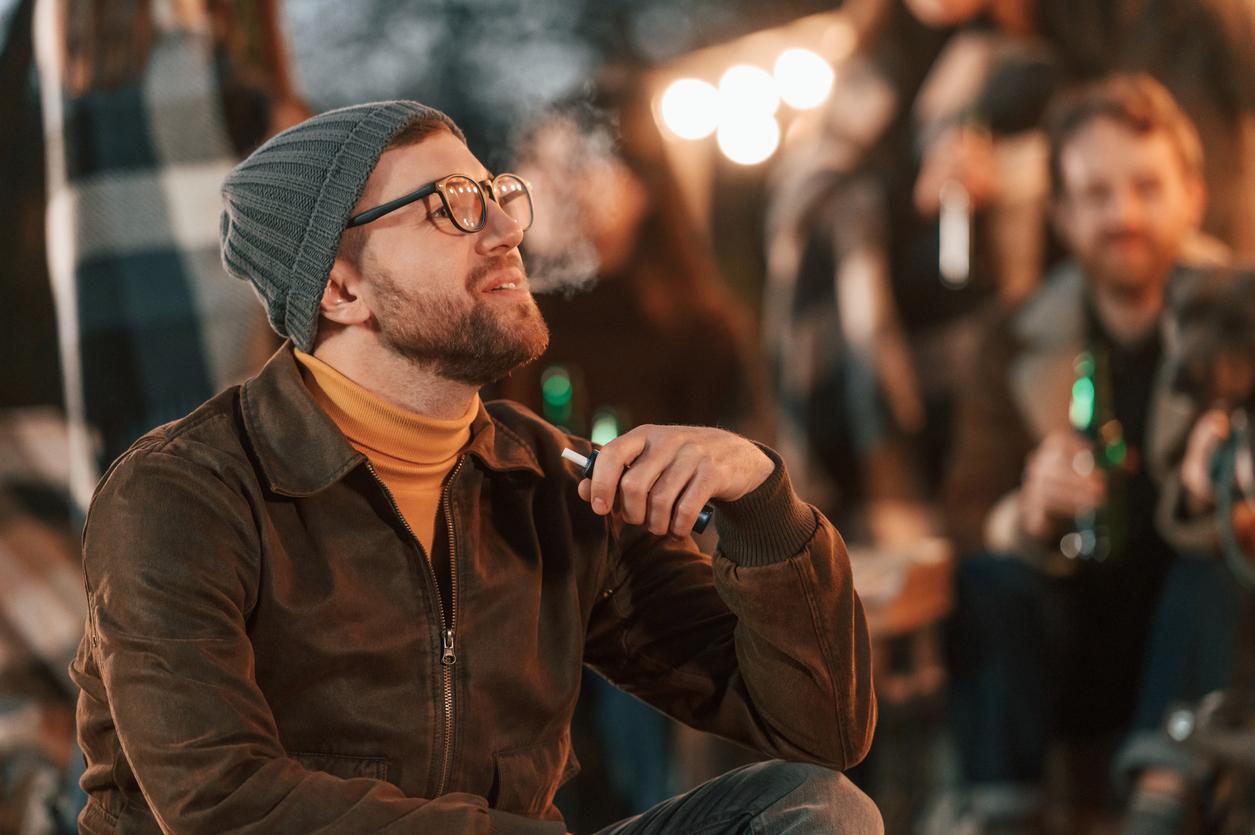 A man wearing glasses and a beanie exhales smoke while holding a vape, sitting outdoors at a social gathering.
