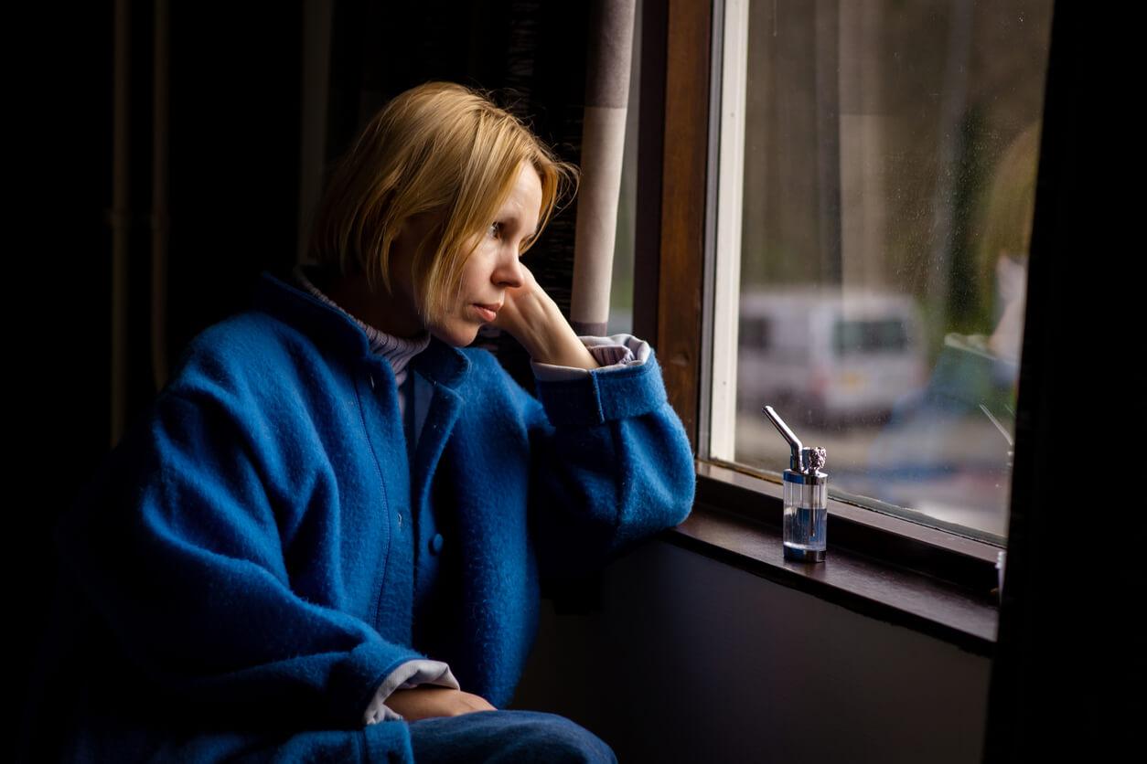 A woman wearing a blue coat sits by a window, looking pensive, with a small vaporiser beside her.