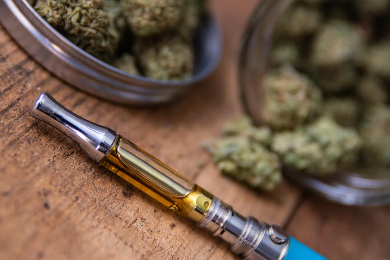 A cannabis vape pen lies on a wooden surface with jars of cannabis buds in the background.