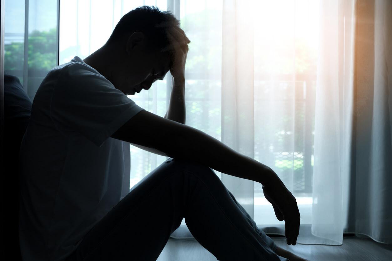 A man sits on the floor with his head in his hand, appearing depressed, in front of a window with sheer curtains.