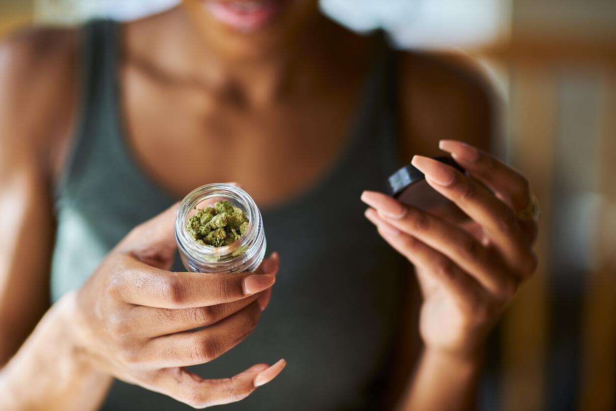 A person inspecting the contents of a small glass jar filled with cannabis buds.