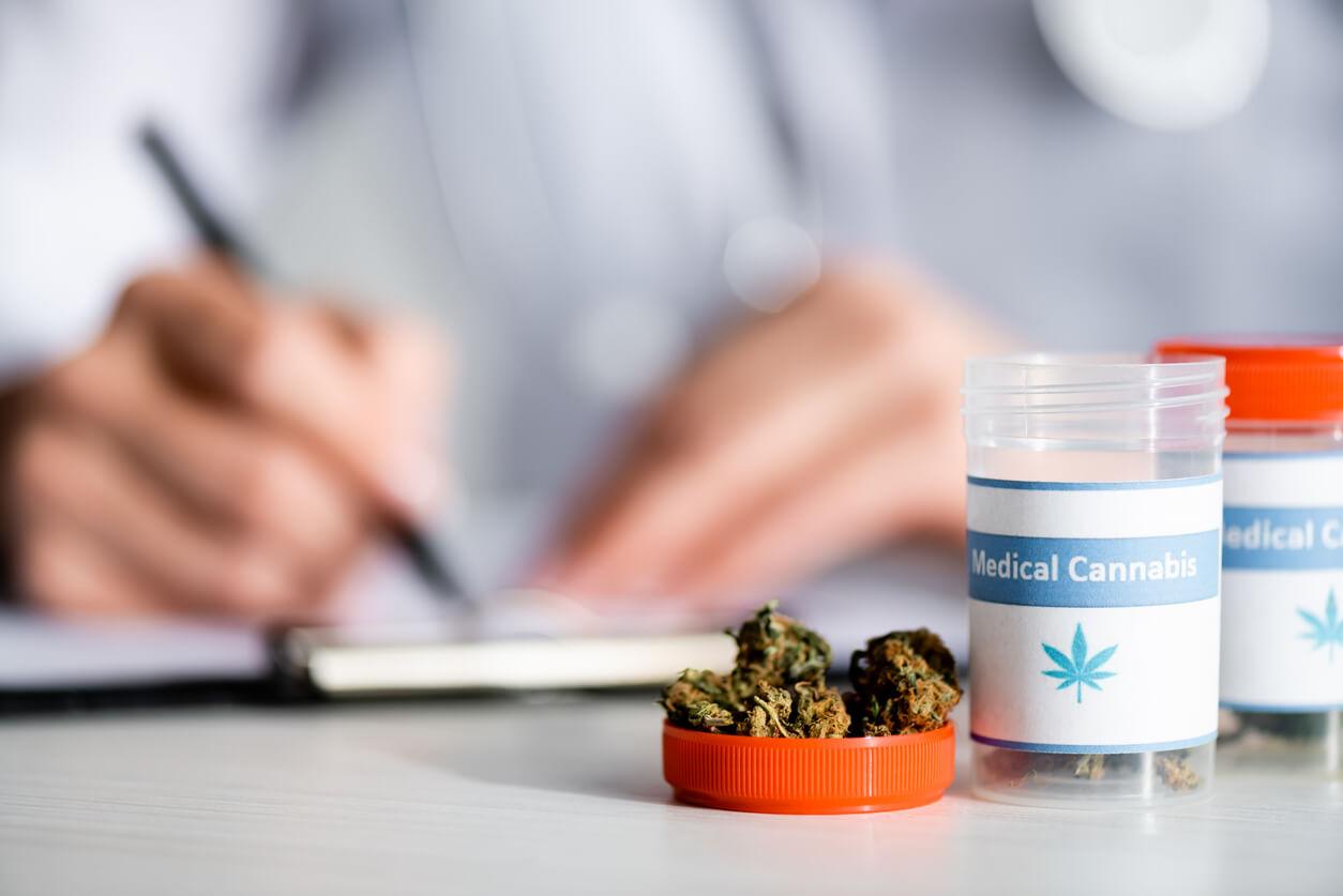 A close-up of medical cannabis containers and buds, with a doctor writing in the background.
