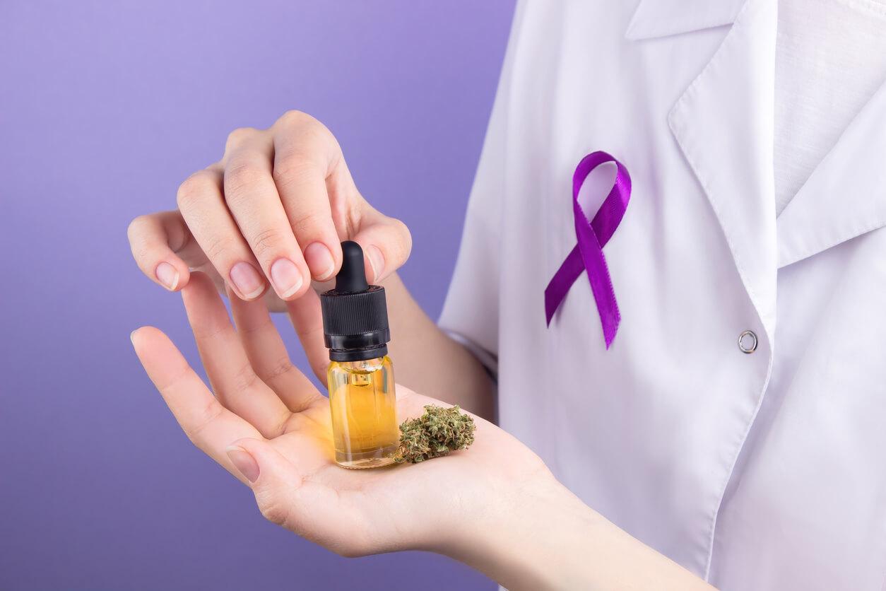 A person in a white lab coat with a purple cancer awareness ribbon is holding a small bottle of cannabis oil and a cannabis bud in their hand. 