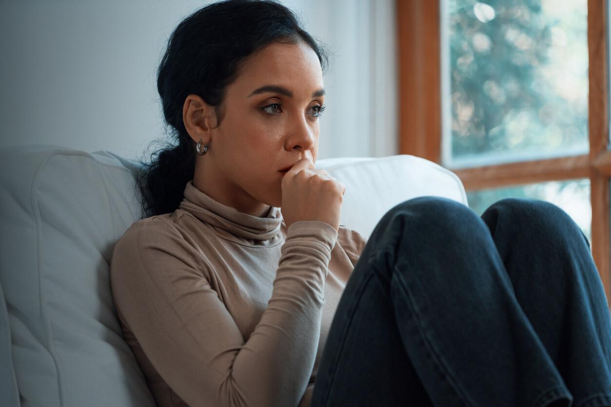 A young woman sits curled up on a sofa, appearing distressed and deep in thought. She is resting her chin on her hand, looking worried and contemplative. The background shows a window with natural light, highlighting the cosy indoor setting and emphasising her emotional turmoil.