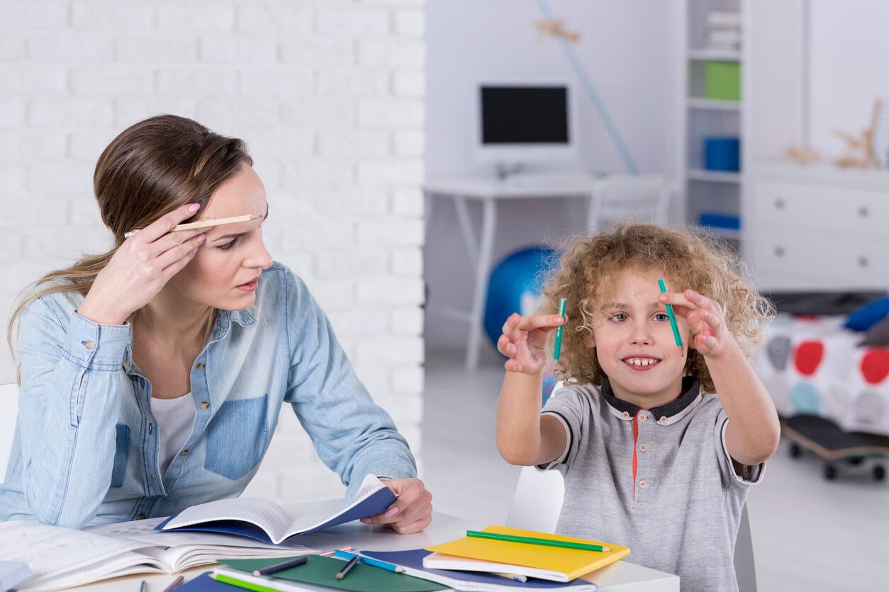 A frustrated woman looks at a book while a smiling, curly-haired child plays with two pencils. They sit at a table with notebooks and stationery in a bright room.
