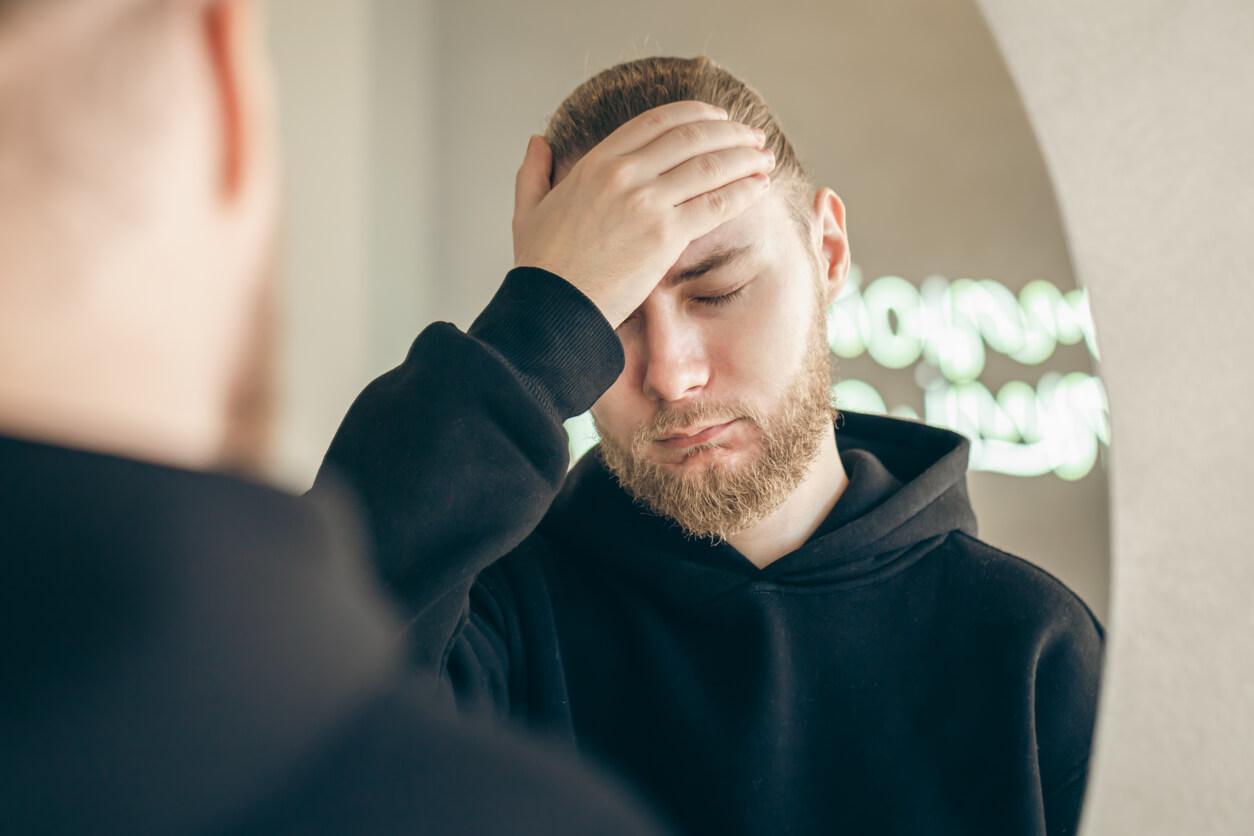 A man with a beard and long hair in a black hoodie is holding his forehead with a pained expression, standing in front of a mirror.