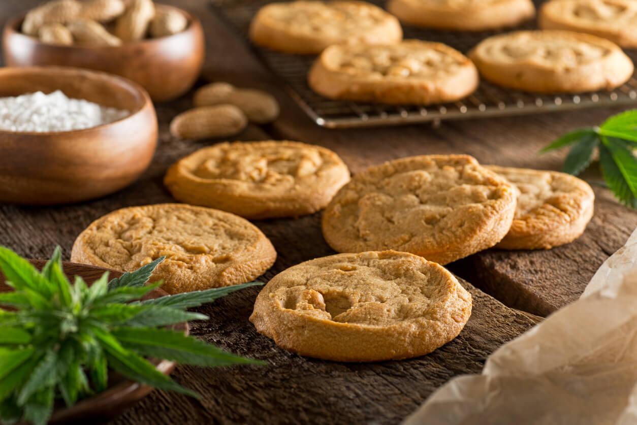 Freshly baked cookies sit on a wooden surface, surrounded by bowls of flour and peanuts, with a cannabis leaf visible in the foreground.