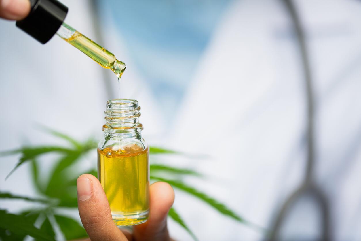A close-up of a hand holding a dropper above a small bottle of yellow cannabis oil, with green marijuana leaves and a person in a lab coat in the background.