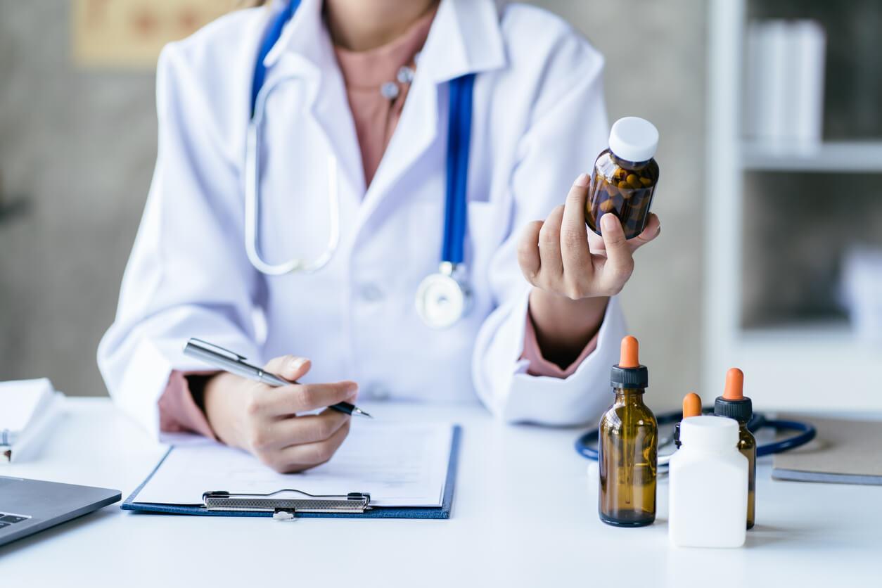 A doctor sits at a desk, holding a bottle of medication while reviewing a clipboard. Several other medicine bottles are on the desk.